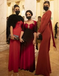 Tre ragazze in abito di gala rosso lungo. Indossano una mascherina.
