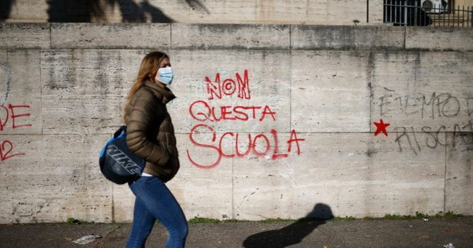 Ragazza con mascherina cammina lungo un muro con un la scritta "No a questa scuola"